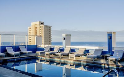 A rooftop swimming pool with lounge chairs overlooks the city and the ocean in the distance under a clear blue sky.