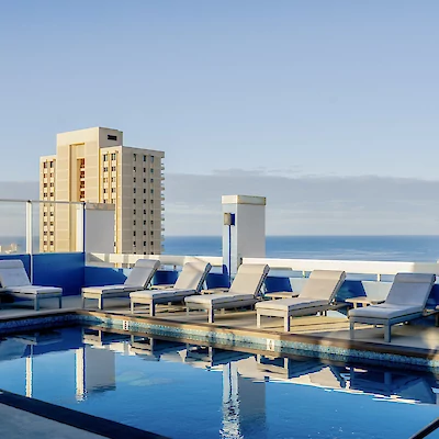 A rooftop swimming pool with lounge chairs overlooks the city and the ocean in the distance under a clear blue sky.