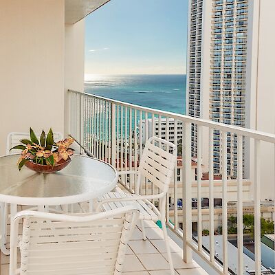 A balcony with a round table, white chairs, and a plant centerpiece, overlooking a cityscape with tall buildings and an ocean view in the distance.