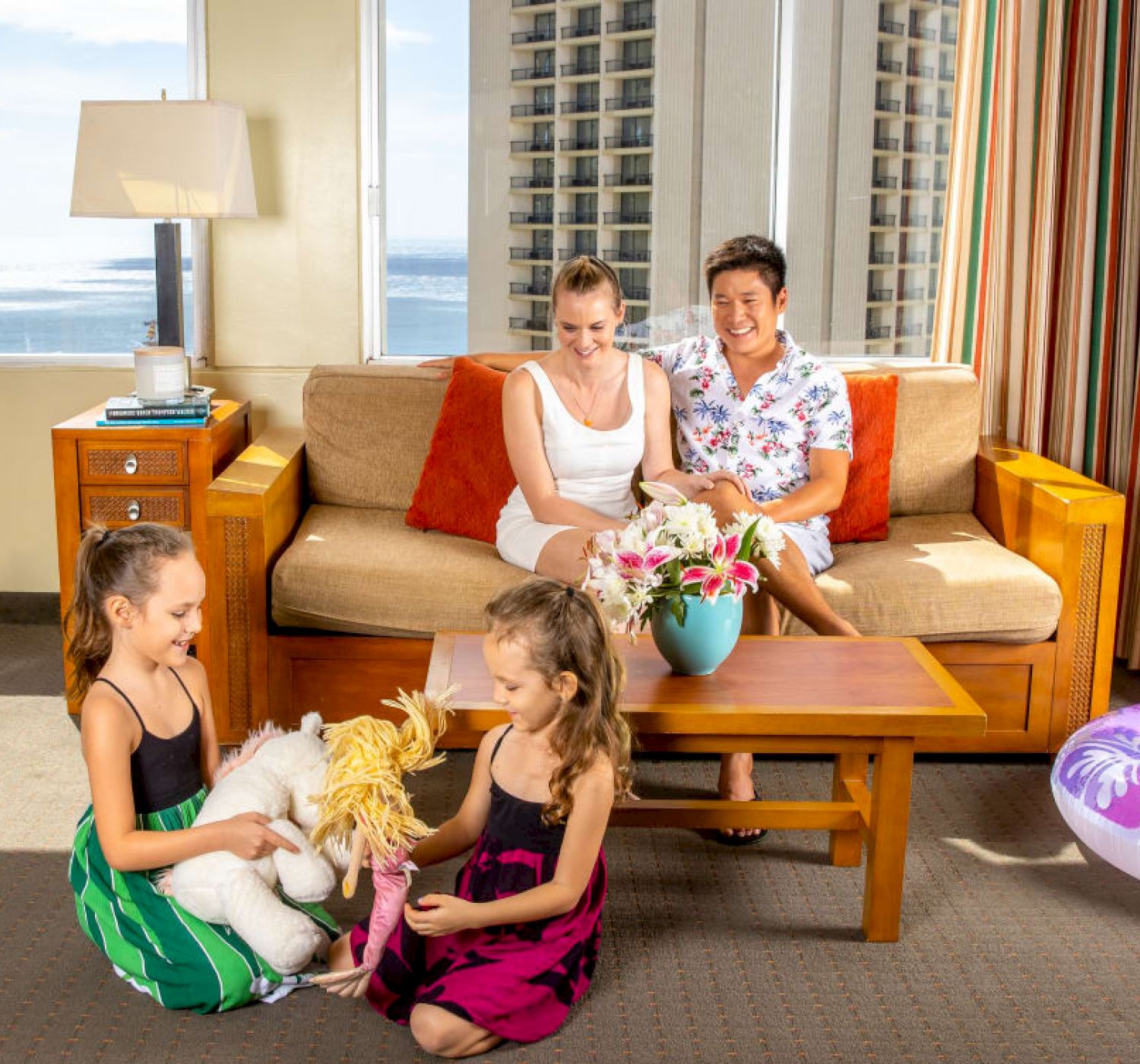 A group of people are in a bright room with a sea view. Two adults are on a couch, while two children sit on the floor playing with toys.