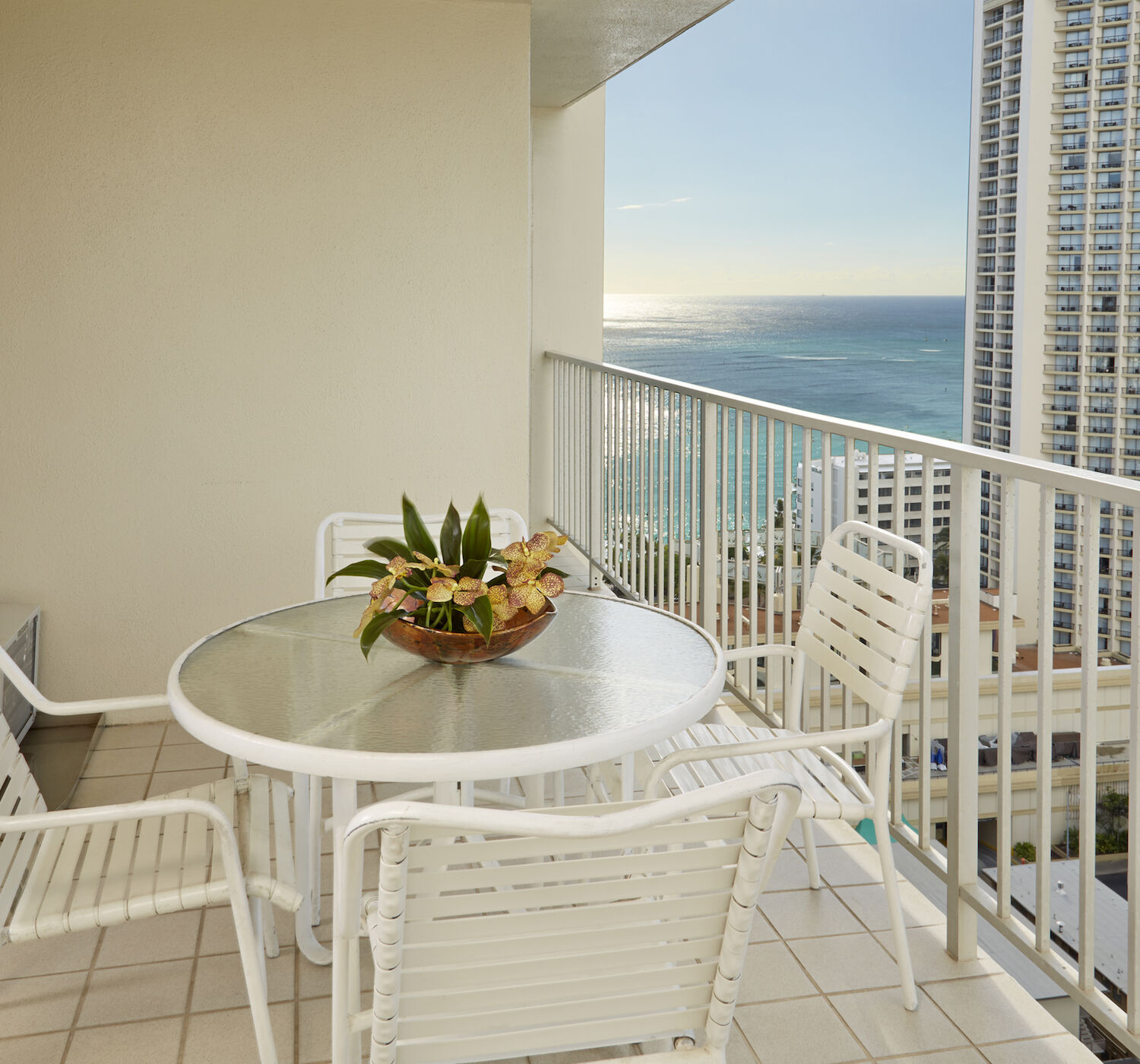 A balcony with a round glass table, four white chairs, and a decorative plant centerpiece overlooks the ocean and tall buildings below.