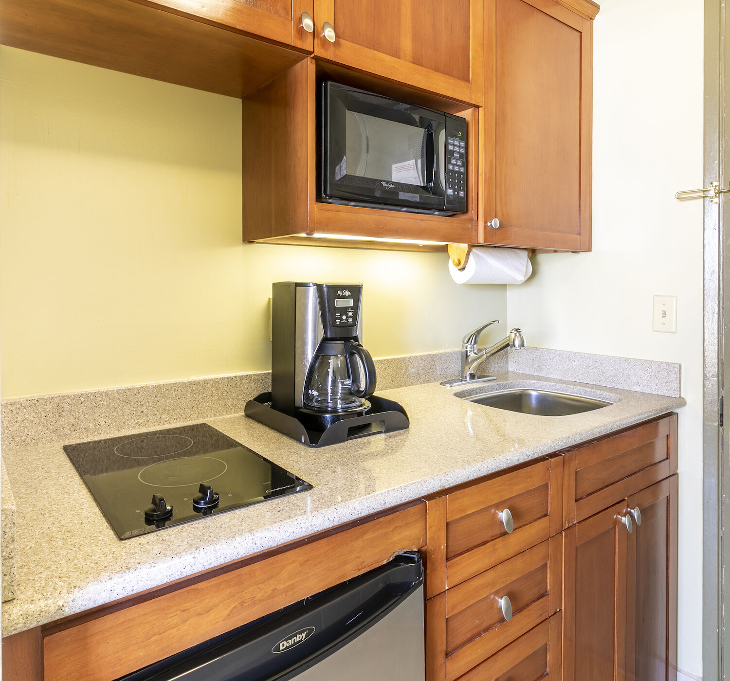 A compact kitchen area with a sink, stovetop, coffee maker, microwave, and a mini-fridge, featuring wooden cabinetry and a beige countertop, is shown.