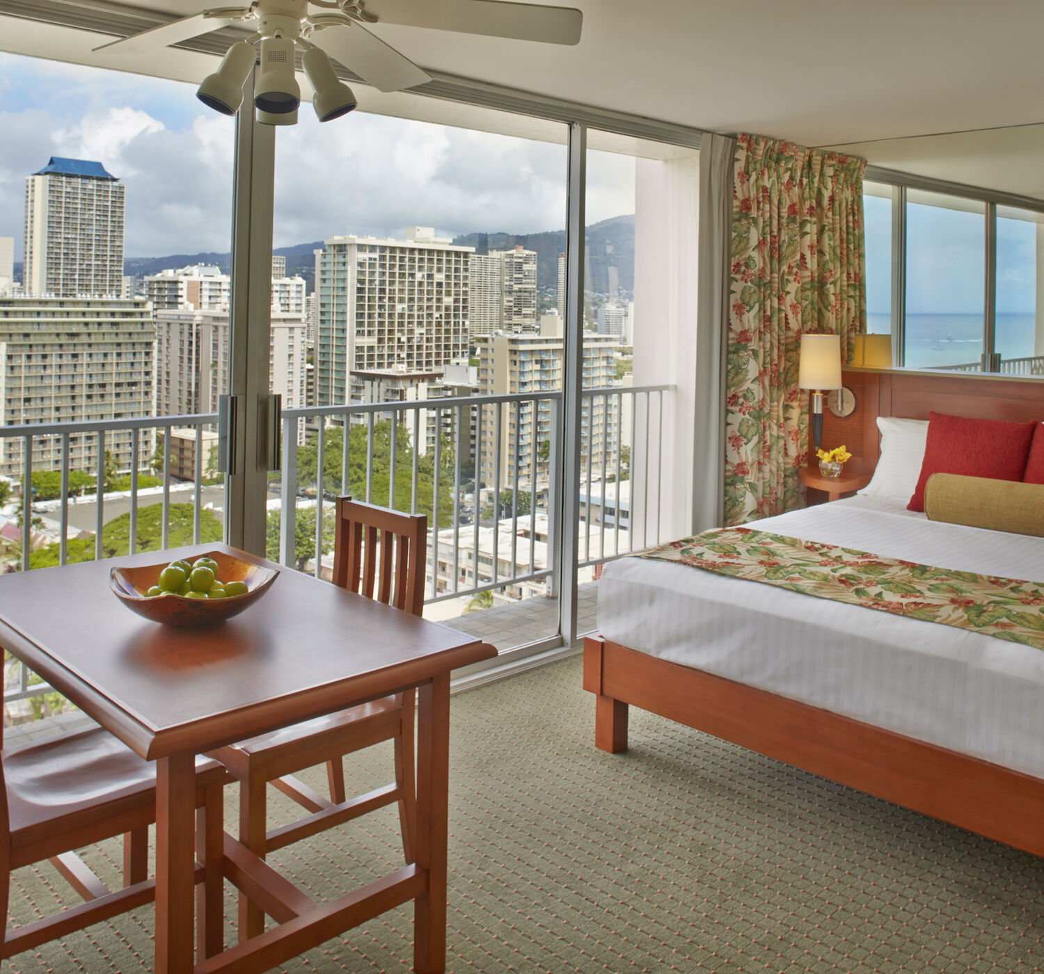 A cozy hotel room with a bed, small dining table, and large windows offering a stunning cityscape view of tall buildings and a glimpse of the ocean.
