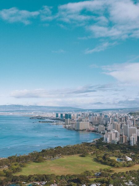 The image shows a coastal cityscape with tall buildings along the shoreline, a sprawling urban area, green fields, and a clear, blue sky.