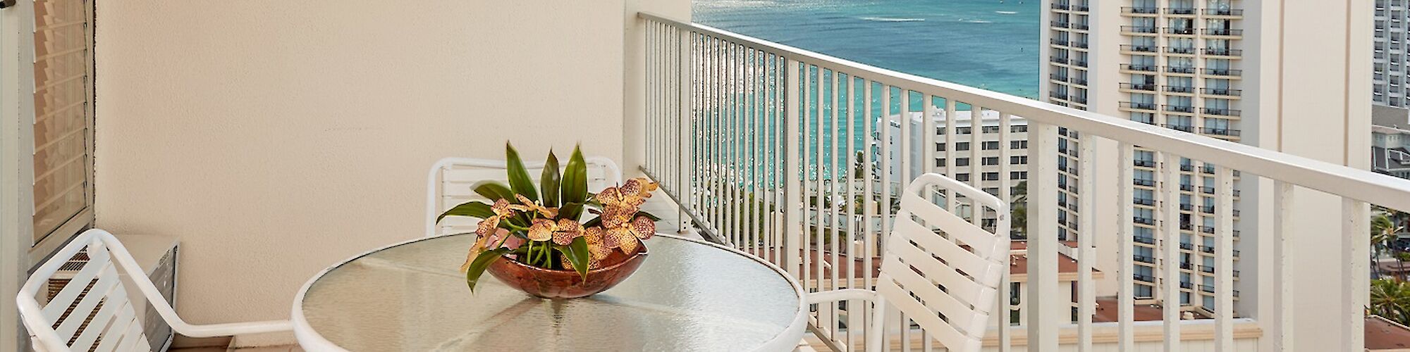 The image shows a balcony with a round glass table, four white chairs, and a potted plant. High-rise buildings and a view of the ocean are visible.