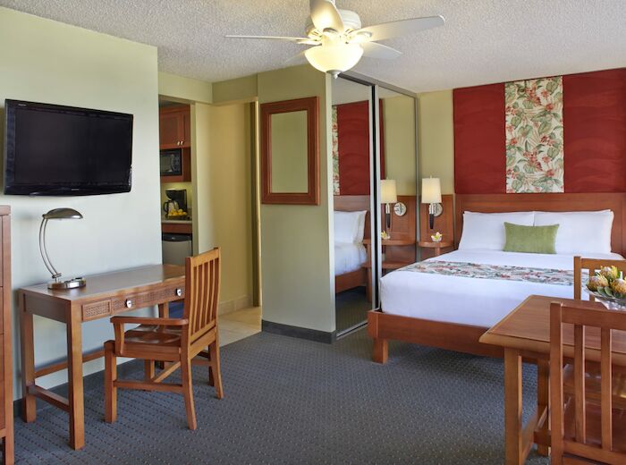 A hotel room with a large bed, desk, TV, ceiling fan, dining table, and a mirrored closet door. A kitchenette is partially visible.