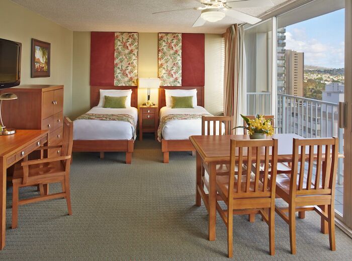 This image shows a hotel room with two beds, a TV, a dining table with chairs, a desk, and a balcony offering a city view.