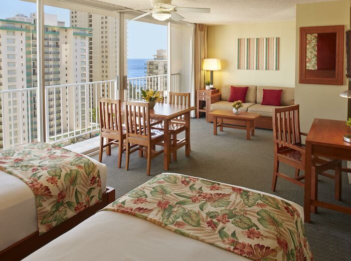 The image shows a hotel room with two beds, a sitting area, a dining table, and a balcony with a view of buildings and the ocean in the distance.