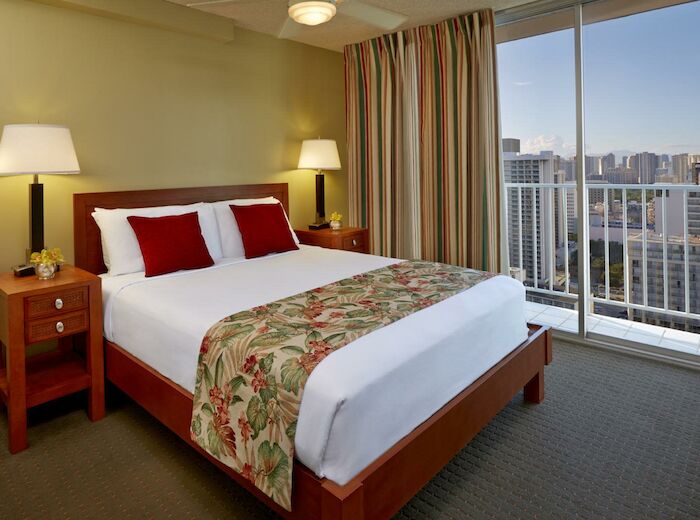 A hotel room with a queen-sized bed, bedside tables with lamps, a colorful bed runner, and a large window with a city view and balcony.