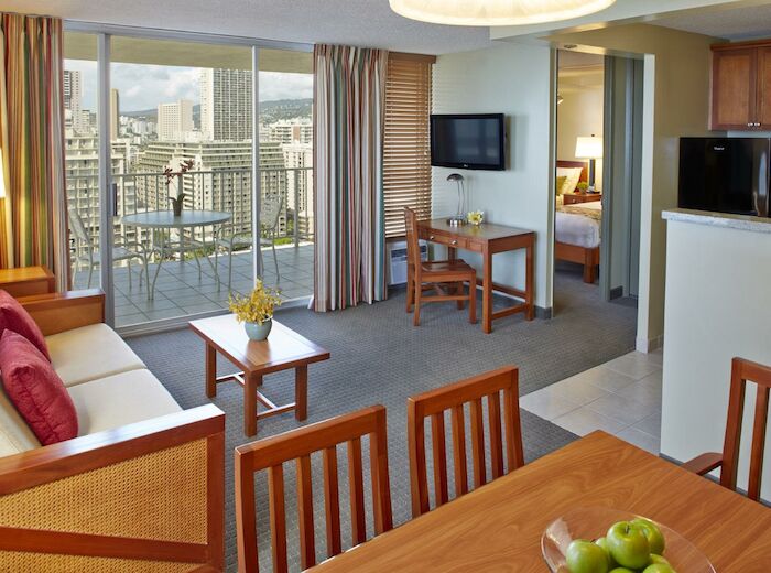 The image shows a hotel suite with a living area, dining table, kitchenette, and a balcony with city views. There is also a separate bedroom visible.