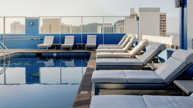 A rooftop swimming pool with multiple lounge chairs lined up next to it. A cityscape is visible in the background under a clear sky.