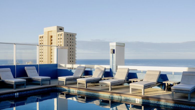The image shows a rooftop pool with several lounge chairs around it, overlooking a cityscape and the ocean under a clear blue sky.