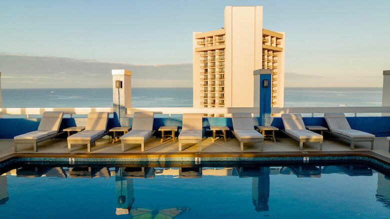 A rooftop pool with lounge chairs beside it overlooks a tall building and the ocean in the distance.