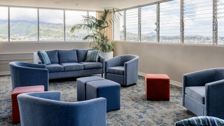 The image shows a sitting area with a blue sofa, blue chairs, and colorful ottomans near large windows with a scenic view outside.
