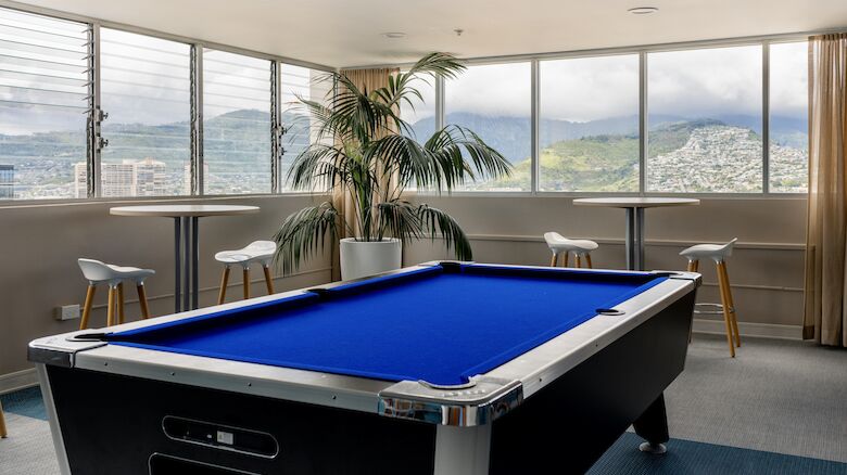 The image shows a room with a pool table, bar stools around high tables, a potted palm, and large windows with mountain views outside.