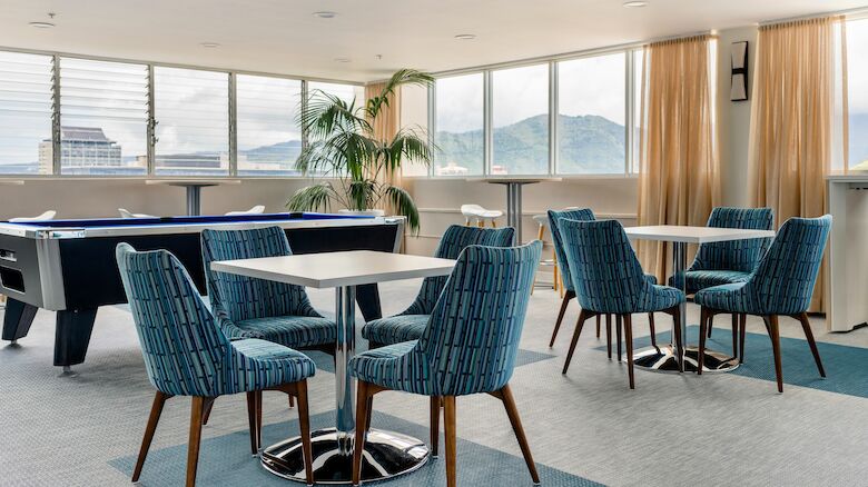 A modern lounge area with tables and blue chairs, a pool table, large windows with view of mountains, and a potted plant in the corner.