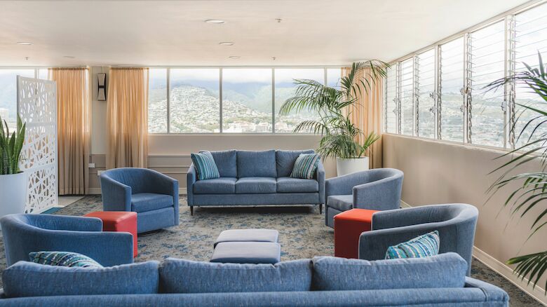 A spacious living room with blue sofas, two red ottomans, large windows, potted plants, and a view of mountains in the distance.