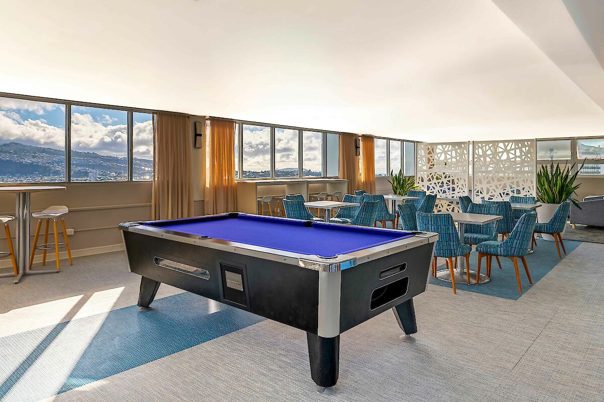 A modern room with a pool table, chairs, tables, and large windows offering mountain views under a bright ceiling.