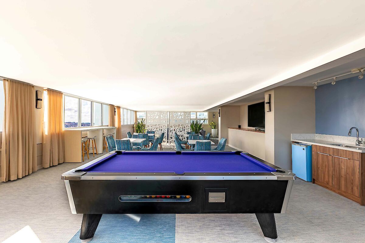 A room with a pool table, seating area, TV, and kitchenette. Bright decor with blue accents and natural light enhance the space.