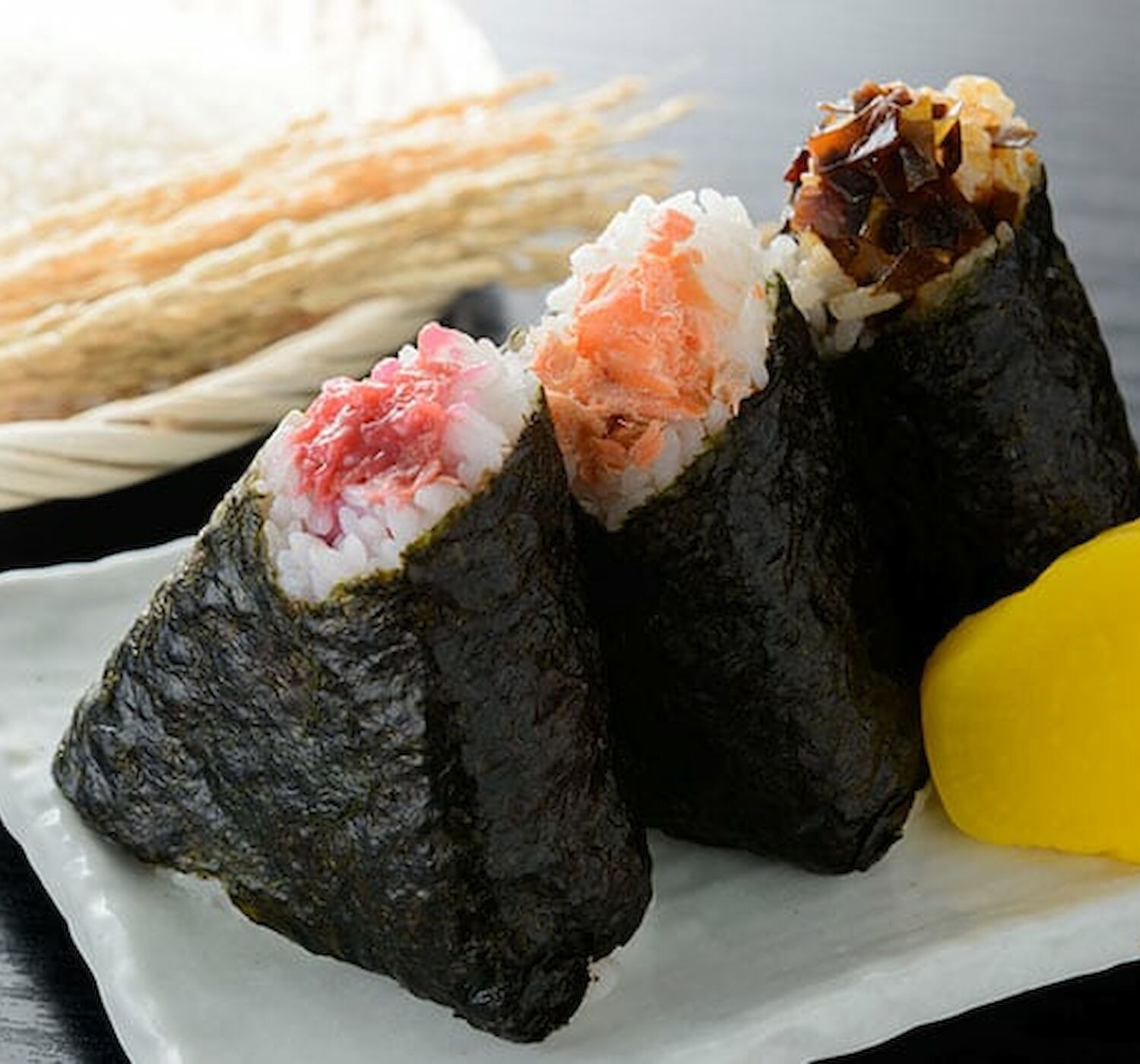 The image shows three onigiri wrapped in seaweed, each with different fillings, served with yellow pickles on a white plate, and a basket in the background.
