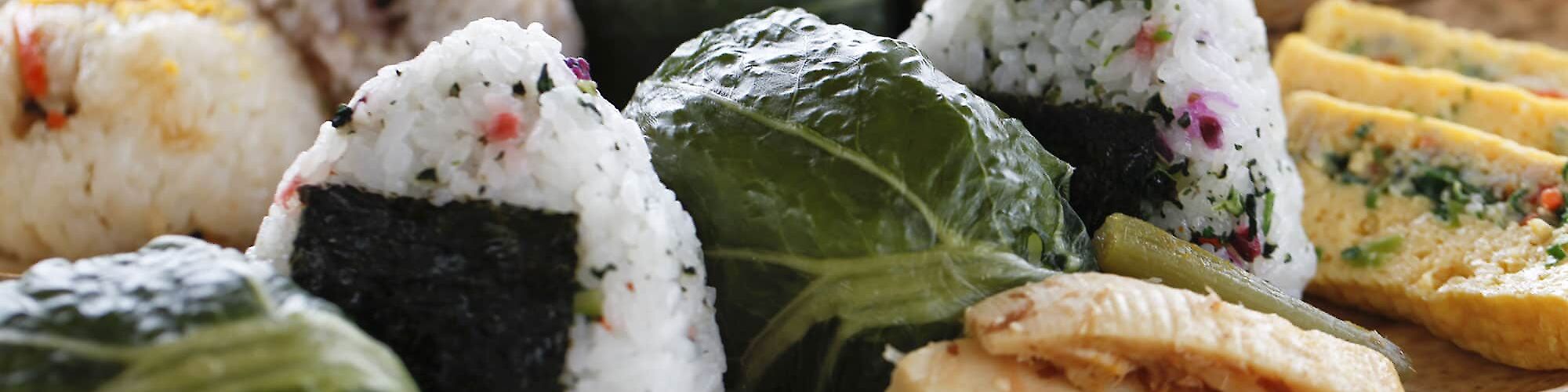 This image features a plate with various types of onigiri (Japanese rice balls) wrapped in seaweed and other leaves, alongside some side dishes.