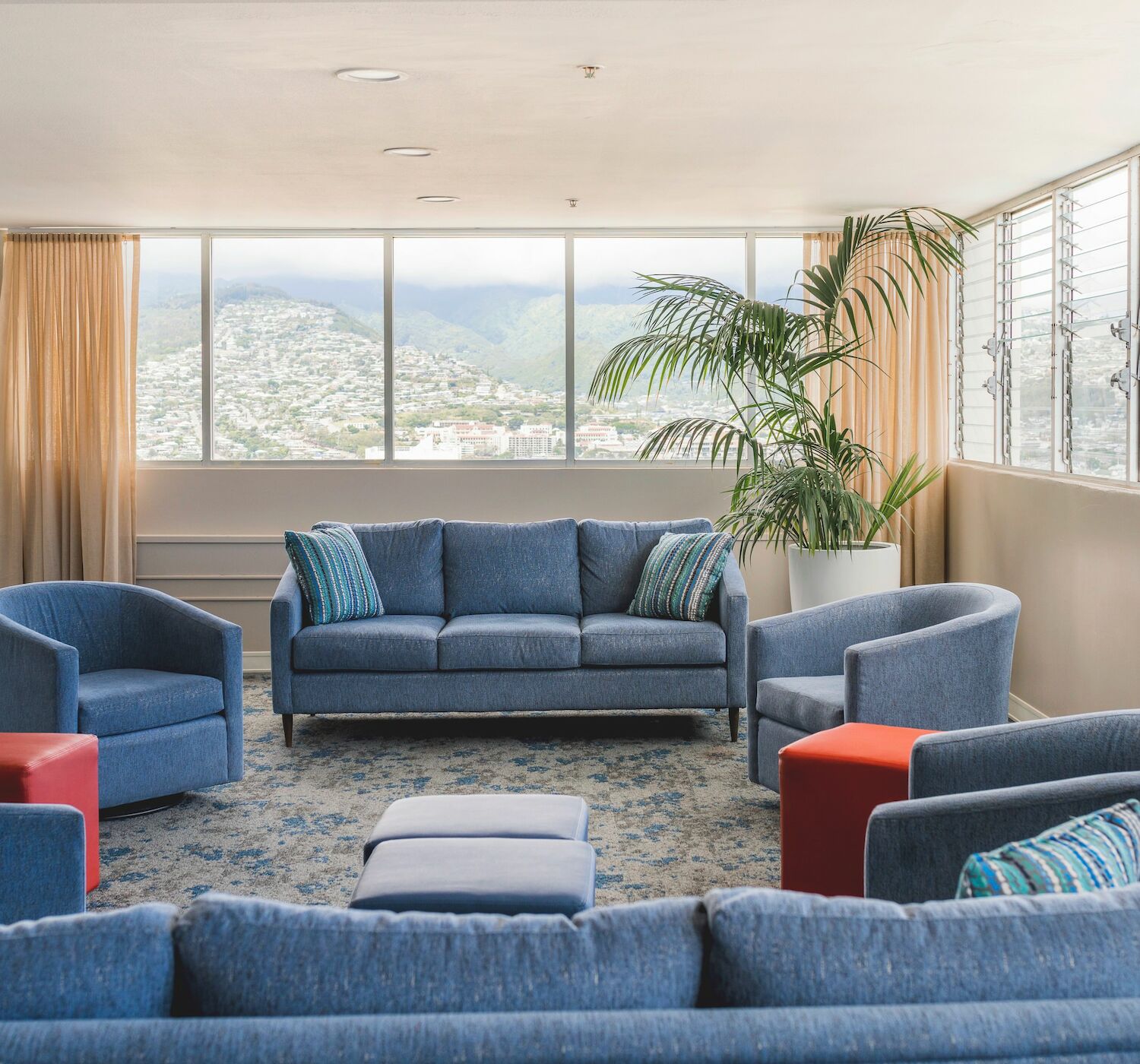 A bright room with blue sofas, orange stools, potted plants, and large windows showcasing a scenic mountain view, creating a relaxing ambiance.