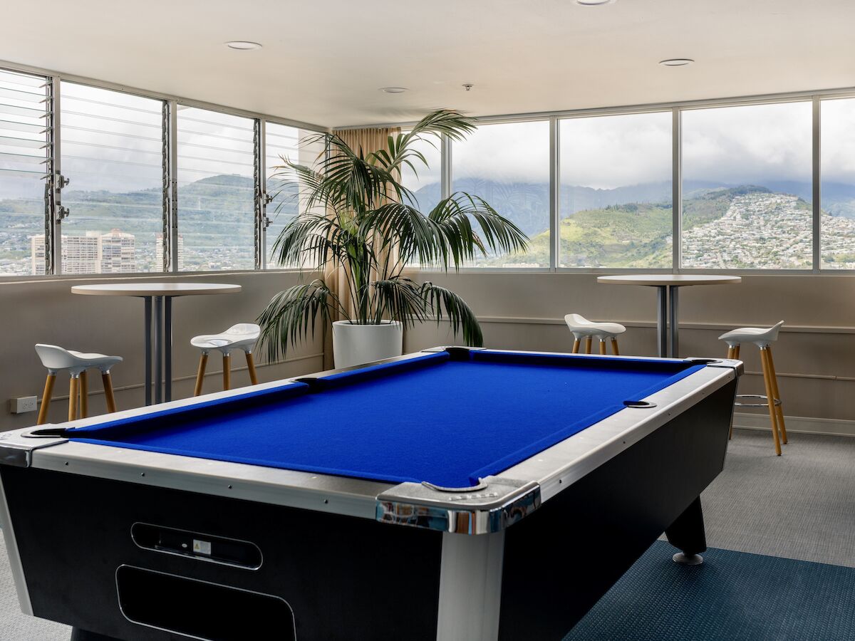 A modern room with large windows offering a mountain view, featuring a blue pool table, bar stools, a tall indoor plant, and tables.