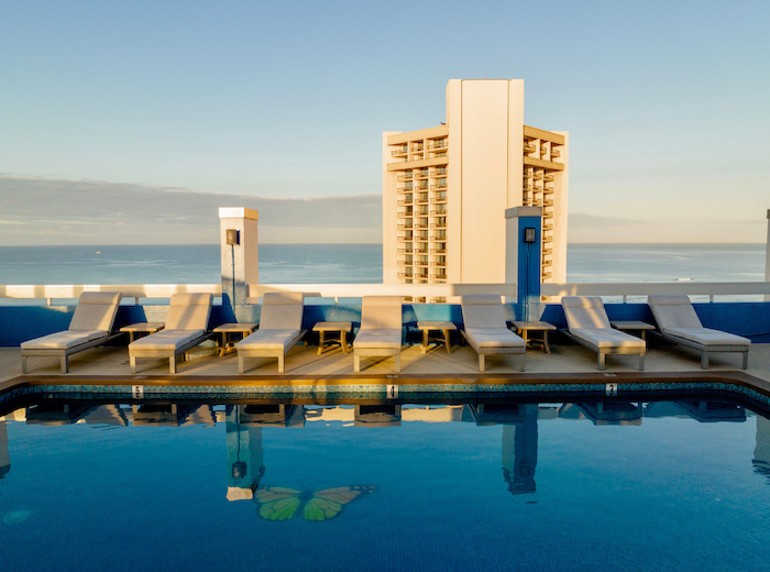 The image shows a rooftop pool with lounge chairs and a view of the ocean and a tall building in the background.