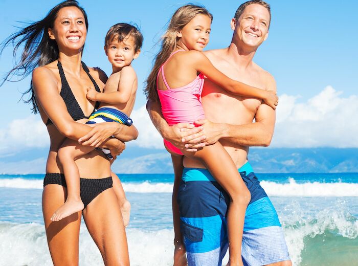 A smiling family of four, in swimwear, is standing at the beach with the ocean waves in the background and clear blue skies overhead.