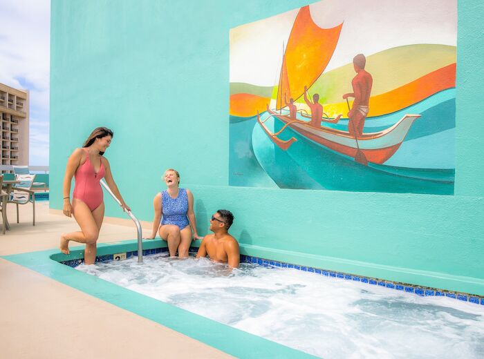 Three people are enjoying a small outdoor pool with a colorful mural of a boat and figures behind them.
