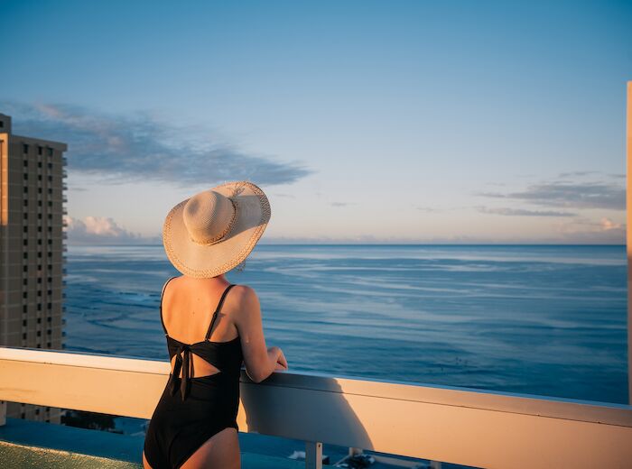 A person in a black swimsuit and hat stands on a balcony overlooking the ocean, flanked by two tall buildings in the background.