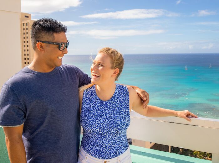A couple is smiling while posing on a balcony with a scenic ocean view in the background. It appears to be a sunny day.