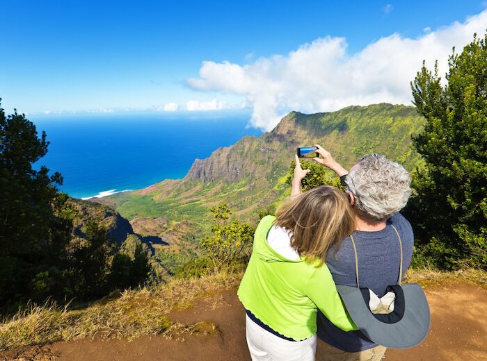 A couple is taking a selfie with a scenic view of mountains and the ocean in the background, surrounded by lush greenery and clear blue skies.