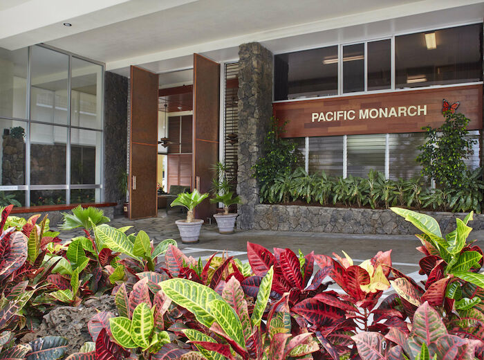 The image shows the entrance to a building named "Pacific Monarch" with large glass doors, stone pillars, and colorful plants in the foreground.