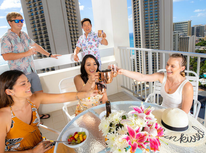 A group of people enjoying drinks on a balcony with a cityscape view, seated at a table with flowers and fruit, smiling and toasting.