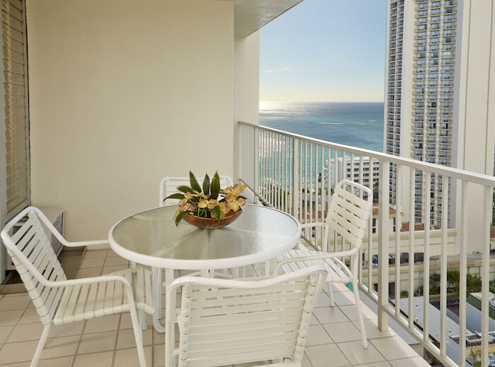 A balcony with white furniture, including a round table and four chairs, overlooks the ocean and a cityscape with tall buildings.