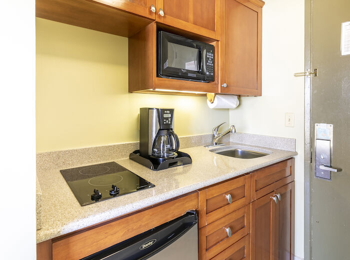 A small kitchen area with a microwave, coffee maker, sink, stovetop, cabinets, and a door with a card reader lock on the right side, ending the sentence.