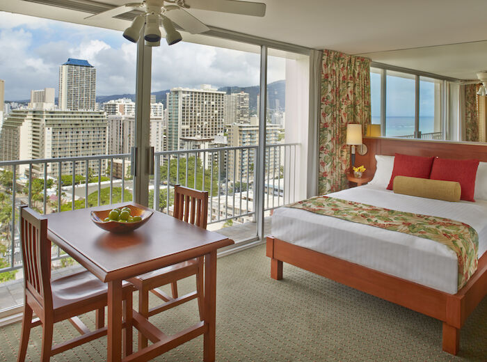 This image shows a hotel room with a wooden table and chairs, a bed with colorful bedding, and large windows overlooking a cityscape.