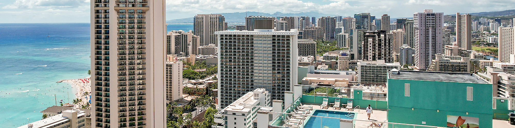 The image shows a skyline with numerous high-rise buildings near a coastline, some of which have rooftop pools, under a clear, blue sky.