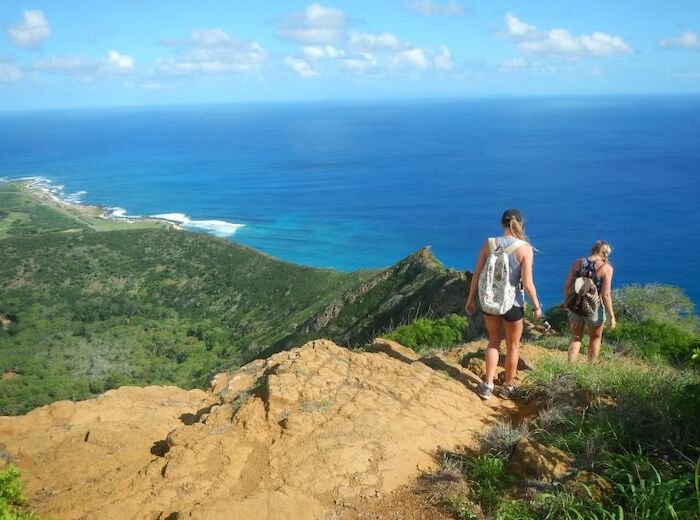 Two people hike down a trail overlooking a scenic coastline with green hills, the ocean, and a partly cloudy sky.