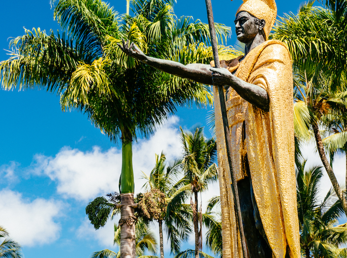 A statue of a person in golden attire stands with arm outstretched, surrounded by tall palm trees under a bright blue sky with some fluffy clouds.