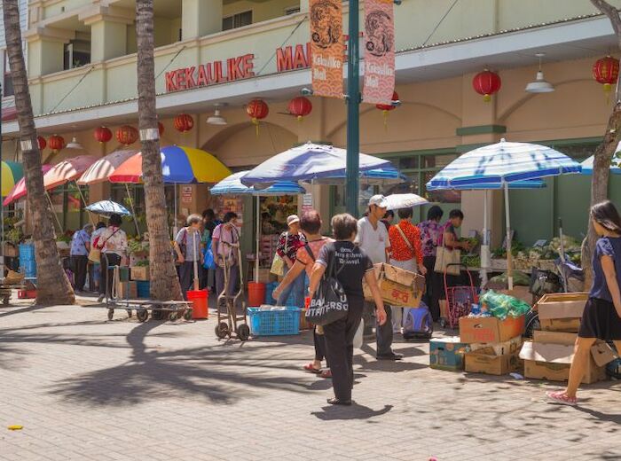 A bustling street market with people shopping, colorful umbrellas providing shade, and a variety of goods displayed outside a store.