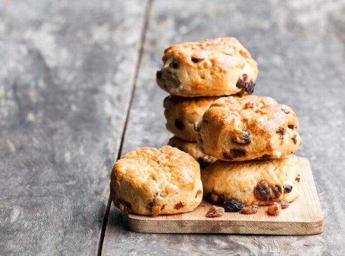 The image shows a stack of rustic-looking scones with raisins on a wooden board, placed on a textured, weathered surface.