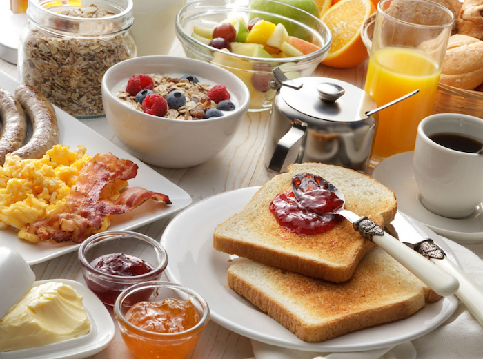 The image shows a breakfast spread with toast and jam, sausages, scrambled eggs, bacon, coffee, juice, cereal, fruit, and various spreads.