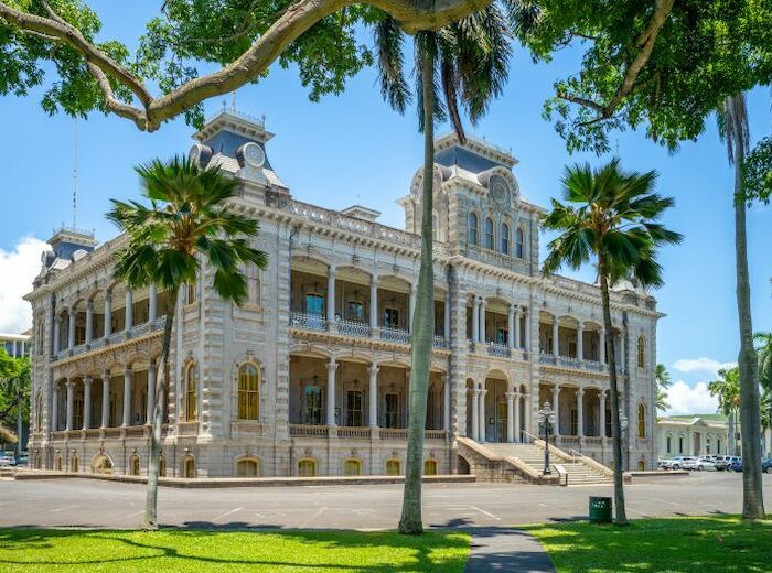 A historic building with ornate architectural details, surrounded by palm trees and a well-maintained lawn on a clear sunny day, is depicted.