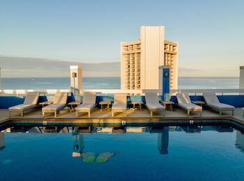 A row of lounge chairs next to a rooftop pool overlooks the ocean with a tall building in the distance under a clear sky.