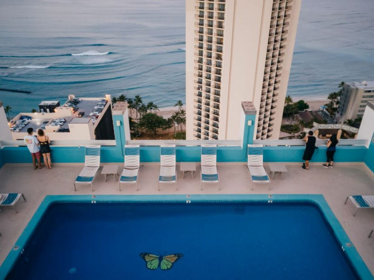 A rooftop pool with a butterfly design at the bottom, surrounded by lounge chairs, overlooks the ocean and buildings.