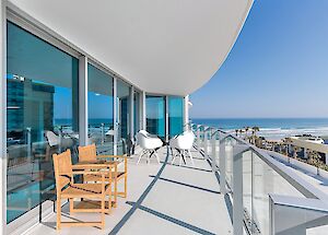 A coastal balcony with wooden and white chairs, glass railing, ocean view, and a modern building nearby on a sunny day.