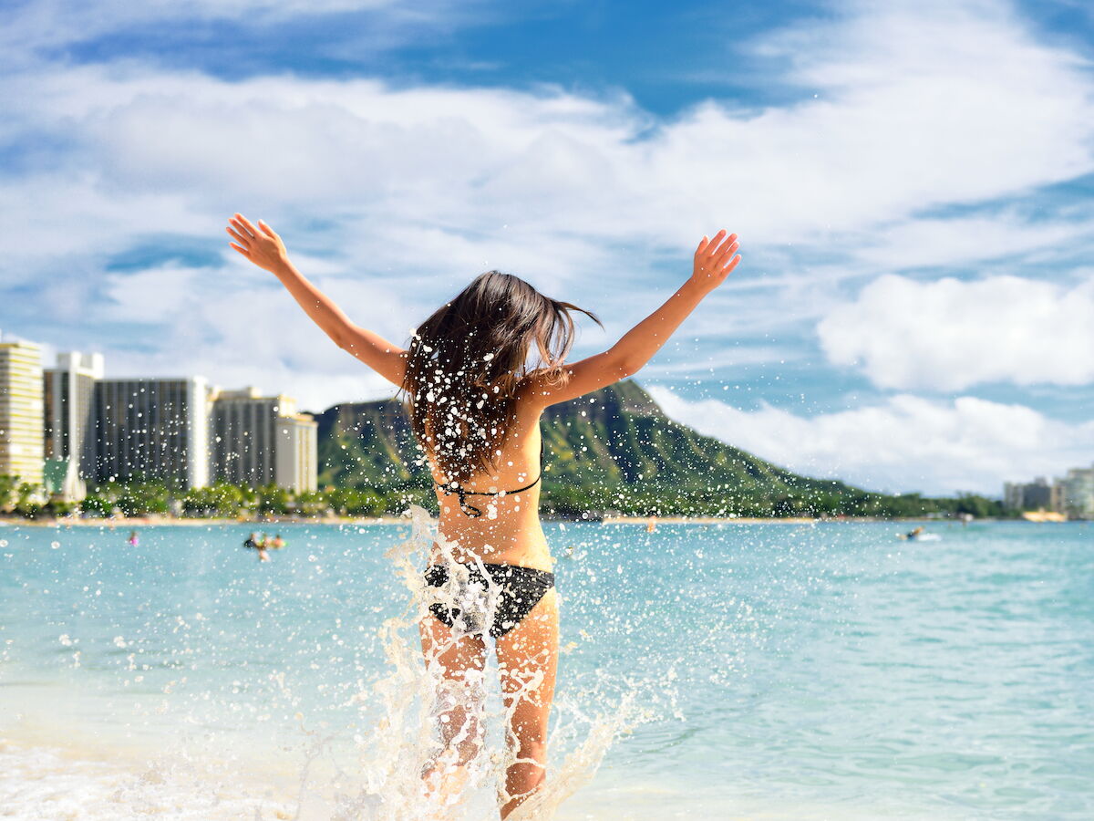 A person in a bikini is joyfully splashing in clear blue water at a scenic beach, with modern buildings and a lush, mountainous backdrop in the distance.