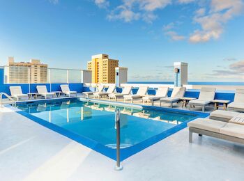 The image shows a rooftop pool with lounge chairs and a view of buildings under a blue sky.